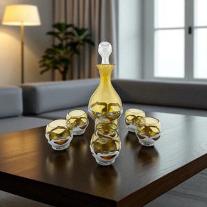 May include: A golden-yellow glass decanter with a clear stopper and a set of matching glasses. The glasses and decanter have silver-colored trim. The set is arranged on a dark brown wooden table.