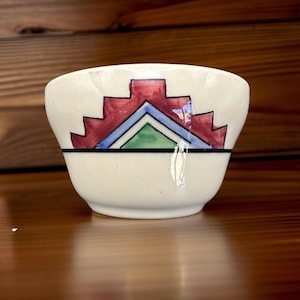 May include: A small, off-white ceramic bowl with a geometric design. The design features a series of red, blue, and green shapes. The bowl has a slightly flared shape and a black band near the rim.