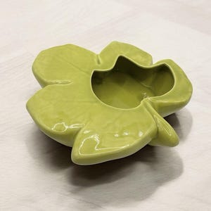 May include: A light green ceramic bowl shaped like a four-petal flower. The bowl has a glossy finish and a central depression. The petals are rounded and slightly curved, with subtle details. The bowl sits on a light-colored surface.