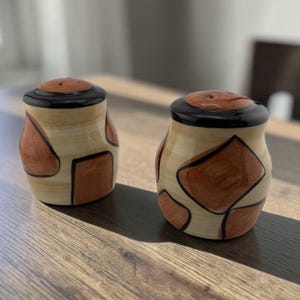 May include: A pair of ceramic salt and pepper shakers with a cream-colored base, featuring hand-painted terracotta-colored geometric shapes outlined in black. Each shaker has a glossy black rim and a terracotta-colored top with small holes.