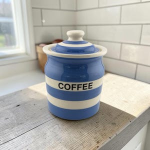May include: A ceramic coffee canister featuring horizontal blue and white stripes. The cylindrical container has a matching lid with a knob handle. The word COFFEE is printed in black capital letters on the center white stripe of the jar.
