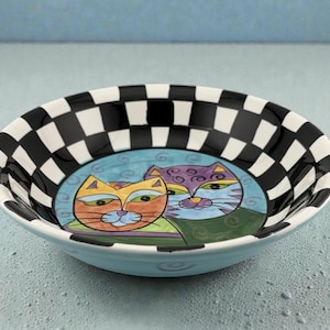 May include: A round ceramic bowl with a light blue exterior and a black and white checkered rim. The interior features a blue background with two stylized cat faces in orange and purple.