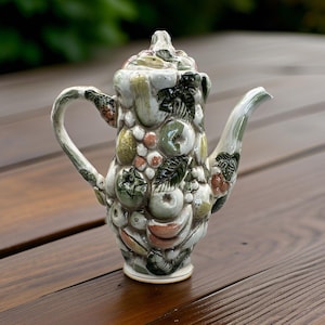 May include: A ceramic teapot with a fruit design. The teapot is white with green, yellow, and red fruits and leaves. It has a curved handle and a spout. The teapot is sitting on a wooden surface.