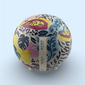 May include: A round ceramic container with a lid, featuring a vibrant tropical floral pattern in pink, yellow, blue, and black. The container has small feet and a label that reads "MEWS COLLECTIVE".