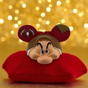 May include: A decorative figurine of Grumpy from Snow White, with Mickey Mouse ears. The figurine is resting on a small, red pillow. The background is a blurred golden color.