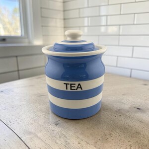 May include: A ceramic tea canister featuring a glossy finish with horizontal blue and white stripes. The cylindrical container has a matching lid with a rounded knob handle. The word TEA is printed in black capital letters on the center stripe.