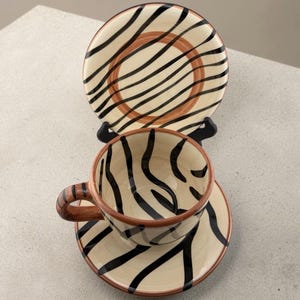 May include: A ceramic teacup and saucer set featuring a cream-colored base with hand-painted black zebra-like stripes and a terracotta-colored rim. The cup has a matching striped handle, and the saucer is displayed upright behind the cup.