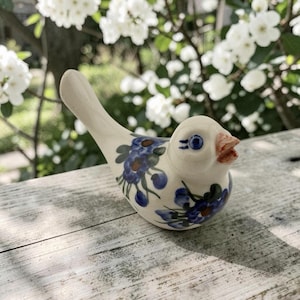 May include: A ceramic bird figurine in an off-white cream color, featuring hand-painted blue floral patterns with green leaves on its body. The bird has a small orange beak, blue eyes with defined lashes, and a long, upward-curving tail feather.