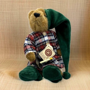 May include: A plush teddy bear wearing a green elf hat and a plaid shirt. The bear has a tag that says "Boyds". The bear is holding a small candle and is sitting down.