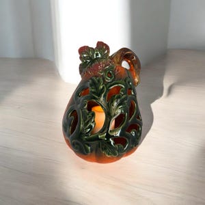 May include: A ceramic pumpkin-shaped candle holder with a handle and leaf details. The pumpkin is orange with green leaf accents and cut-out designs. A lit candle is visible inside the holder, casting a warm glow.