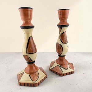 May include: Two ceramic candle holders featuring a glossy finish with a geometric pattern in shades of cream, terracotta brown, and black. Each holder has a flared top, a slender neck, and a hexagonal base with black vertical line accents.