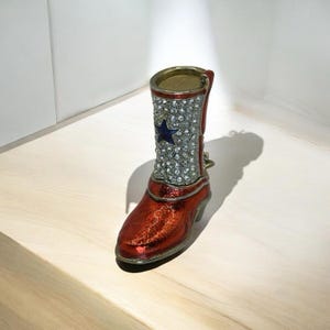 May include: A decorative red and silver cowboy boot with a blue star on the shaft. The boot is embellished with rhinestones and has a metallic finish. The boot is a small decorative item.