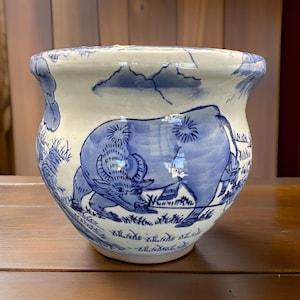 May include: A ceramic planter with a wide, slightly flared rim. The planter is white with blue illustrations of a water buffalo, grass, and clouds. The design is hand-painted, giving it a unique, artistic look. The planter is suitable for indoor or outdoor use.