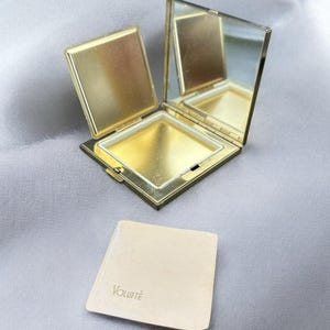 May include: A vintage gold-toned compact mirror, open to reveal a mirror and powder compartment. A small, square powder puff with the word "Volupté" is next to the compact. The compact and puff rest on a gray, silky surface.