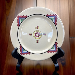 May include: Cream-colored ceramic plate with a black rim and a central design featuring a yellow circle with red and blue accents. The plate has a geometric pattern in red, blue, and green at the corners.