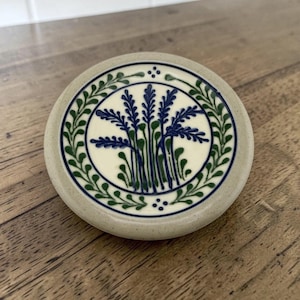 May include: A round ceramic lid with a speckled light gray base. The center features a hand-painted design of blue and green wheat stalks enclosed in a circular border. A decorative green vine pattern encircles the central motif on a cream background.