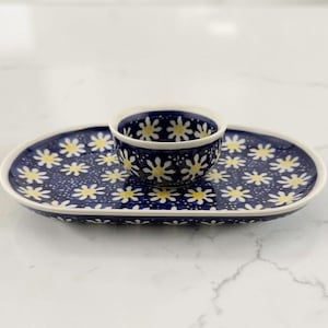May include: A ceramic oval tray with a small matching bowl centered on top. Both pieces feature a dark blue background with a repeating pattern of white daisy flowers with yellow centers. The edges of the tray and the rim of the bowl are white.