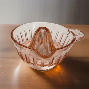 May include: A vintage pink glass citrus juicer with a reamer and a handle. The juicer has a ribbed design on the bowl and reamer. The glass is translucent, and the juicer is sitting on a wooden surface.