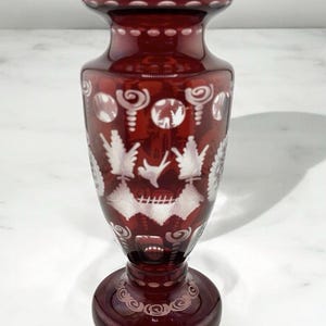May include: A ruby red glass vase with a wide base and flared top. The vase features intricate white etched designs, including floral and geometric patterns. The vase is approximately 10 inches tall.