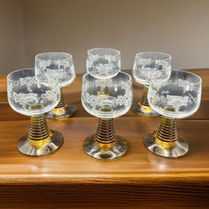 May include: Six clear wine glasses with etched grape leaf designs. The stems have a gold-colored, ribbed design, and the bases are gold. The glasses are arranged on a wooden surface.