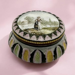 May include: A decorative, round trinket box with a lid featuring a painted scene of a person near a body of water. The box has a light gray base with green, yellow, and pink accents. The lid is trimmed with pink and green.