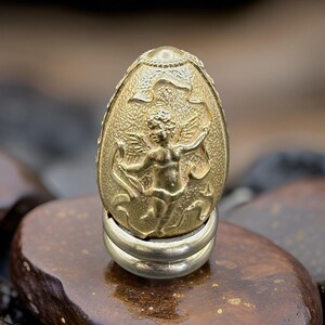 May include: A gold-colored, egg-shaped decorative object with an embossed design featuring a cherub with wings. The egg sits on a silver-colored base. The background is blurred, with a focus on the ornate details of the egg.