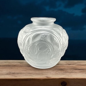 May include: Frosted glass vase with a round body and a small, cylindrical neck. The vase features a detailed relief pattern of leaves and berries. The design is symmetrical and wraps around the entire body of the vase. The vase is sitting on a wooden surface.