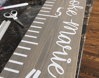 May include: A gray wooden growth chart with white markings and the name "Emily Brooke" written in white cursive lettering.