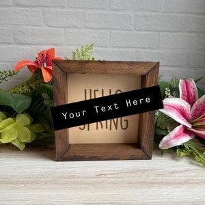May include: A wooden sign with the text "Hello Spring" and a blank space for personalized text. The sign is brown and has a light wood background. The sign is surrounded by green and pink flowers.