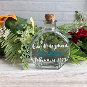Pode incluir: Uma garrafa de vidro transparente com uma rolha de cortiça. A garrafa tem letras brancas que dizem "Our Honeymoon St. John February 2023".