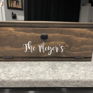 May include: A wooden box with a hinged lid and a black knob. The box is stained a dark brown colour and has the text "The Meyer's" written in white on the front.