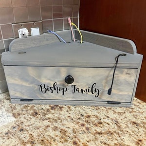 May include: A gray wooden box with a hinged lid and a small black knob. The box has a cutout on the top for cords and is labeled "Bishop Family".