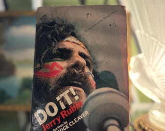May include: Vintage book cover for "Do It!" by Jerry Rubin, featuring a portrait of a man with red face paint. The title is in bold white letters with a red accent. The book is displayed on a black stand.