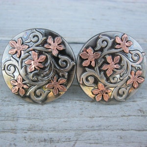 May include: A pair of silver and copper floral earrings with a detailed design. The earrings are round and have a floral pattern with copper accents.