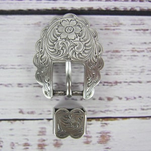 May include: Silver-toned belt buckle with an ornate floral design. The buckle is attached to a matching silver-toned strap keeper.