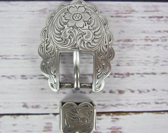 May include: Silver-toned belt buckle with an ornate floral design. The buckle is attached to a matching silver-toned strap keeper.