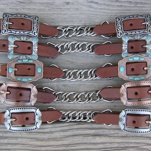 May include: Five brown leather straps arranged horizontally, each featuring two metal buckles connected by a central silver-toned chain. The buckles display various engraved patterns and some are inlaid with small, round, light blue turquoise stones.