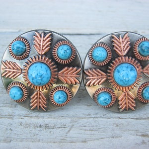 May include: A pair of round, silver and copper concho buttons with turquoise stones and a geometric design.