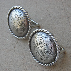 May include: A pair of silver-toned cufflinks with a rope-like border and a floral design.