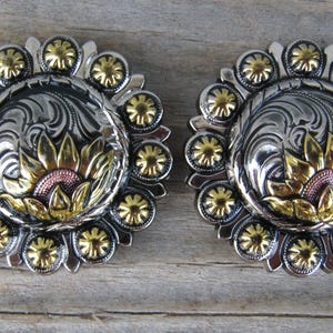 May include: Two silver and gold-tone metal conchos with a floral design. Each concho features a central sunflower design with gold petals and a silver filigree background. The outer edge has gold accents.