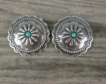 May include: Two silver-toned, round concho buttons with a daisy design and a turquoise center. The buttons have a rope-like border and are set against a weathered wood background. These decorative buttons are suitable for various craft projects.