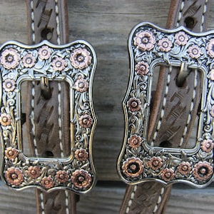 May include: Two silver-toned buckles with copper-colored floral accents. The buckles are rectangular with ornate floral designs and are attached to brown leather straps with white stitching. The background is a wooden surface.