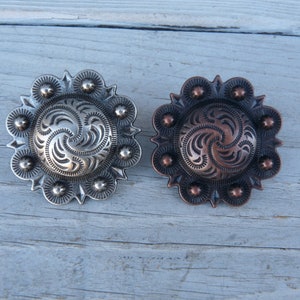 May include: Two decorative metal conchos with a swirl design. The concho on the left is silver and the concho on the right is copper.