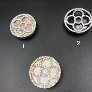 May include: Three round, decorative phone grips with a flower-like design. The first grip has a white and iridescent shell background, the second has a black background, and the third has a white background. All three grips are surrounded by a ring of clear rhinestones.