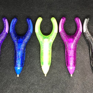 May include: Six colorful glitter pens with a unique ergonomic design. The pens have a clear plastic body with glitter embedded in the plastic. The pens are in the colors purple, blue, green, pink, gray, and black.