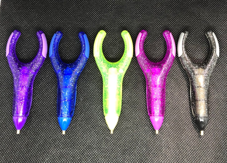May include: Six colorful glitter pens with a unique ergonomic design. The pens have a clear plastic body with glitter embedded in the plastic. The pens are in the colors purple, blue, green, pink, gray, and black.