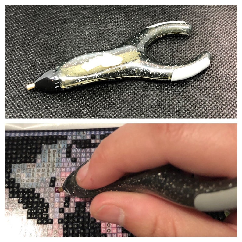 May include: A close-up of a black and silver diamond painting pen with a clear tip. The pen is being used to place a diamond on a diamond painting canvas.
