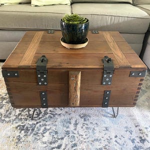 Op de afbeelding: Een rustieke houten salontafel met metalen accenten en haarspeldpoten. De tafel is gemaakt van gerecycled hout, met zichtbare nerven en metalen scharnieren. Bovenop staat een donkerblauwe keramische pot met een vetplant. De tafel meet ongeveer 76 cm vierkant.