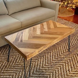 May include: A rectangular coffee table with a herringbone pattern made of light-coloured wood. The table has black hairpin legs and is set on a patterned rug. The table is near a beige sofa.