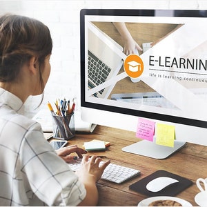 May include: A woman sits at a desk in front of a computer monitor. The monitor displays a graphic with the text "E-LEARNING life is learning continuously".